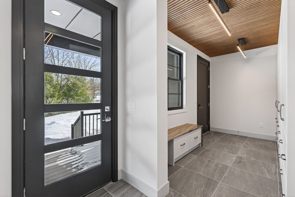 Custom Maine mudroom featuring a black glass exterior door, built-in stone bench, white storage cabinetry, gray tile flooring, and wood slat ceiling.