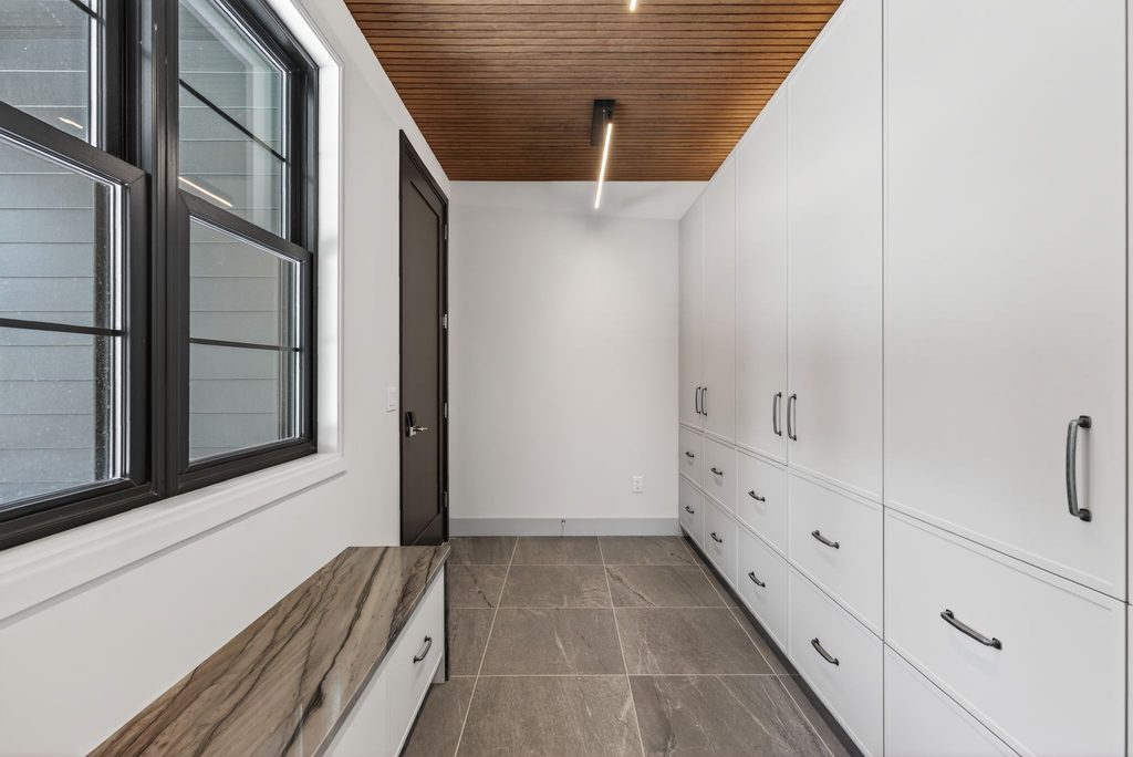 Custom Maine mudroom featuring floor-to-ceiling white cabinetry, a built-in stone bench, large black-framed windows, and durable tile flooring.
