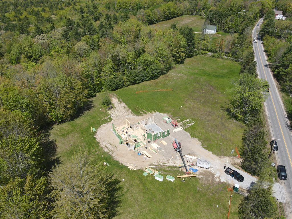 Aerial photograph of a custom home under construction on a cleared Maine property, showing wood framing, construction equipment, and site access.